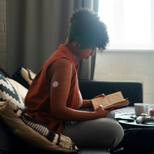 A Black woman wearing a glucose sensor sits forward on a couch, reading a book with interest